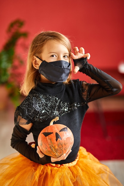 Girl at home in halloween costume with pumkin Jack or Laurent in hands, Child wearing black face mask protecting from coronavirus, halloween in quarantine