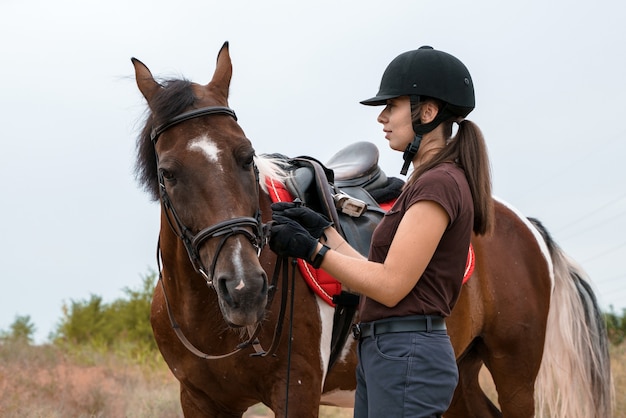 Una ragazza in attrezzatura per l'equitazione si trova su una strada di campo accanto al suo cavallo pezzato e lo tiene per le redini