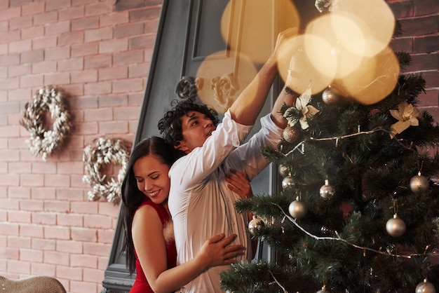 Girl hugging her boyfriend with love. Romantic couple dressing up Christmas tree in the room with brown wall and fireplace.