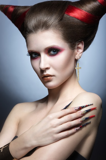 The girl in an image of the demon-tempter with long nails and haircut in the form of horns. Picture taken in the studio on a gray background.