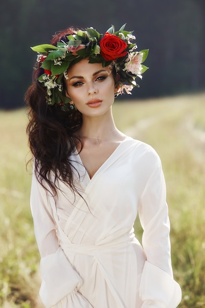 Фото Девушка в длинном платье стоит в поле с венком на голове и букетом цветов в руках, красивая женщина в лучах вечернего солнца осенью в деревне