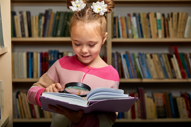 Фото Девочка в повседневной одежде в библиотеке после школы, читает энциклопедию, прилежный ребенок использует увеличительное стекло