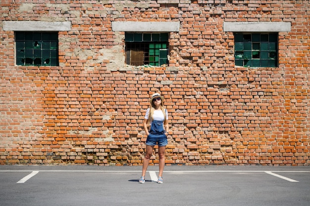사진 밝은 태양 아래 벽돌 건물 배경에 데님 작업복을 입은 소녀, 흰색 티셔츠, 가벼운 모자