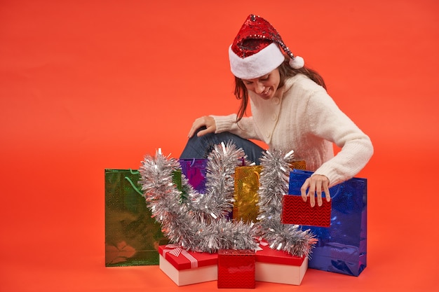 Фото Девушка в шляпе санта-клауса с радостью размещает рождественские подарки с сумками и украшениями на оранжевом фоне