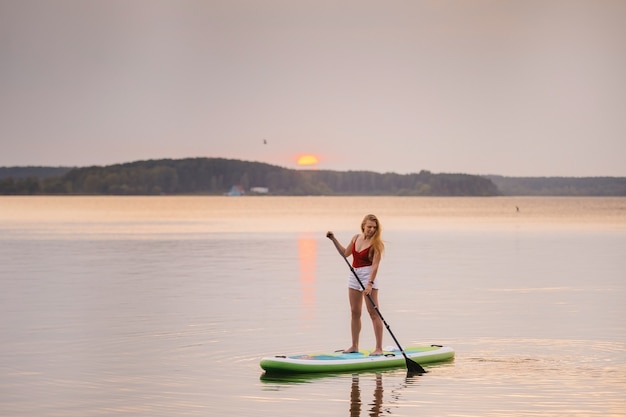 写真 海の少女は、美しい夕日や日の出の水の色でパドルsupボードの上に立ちます。