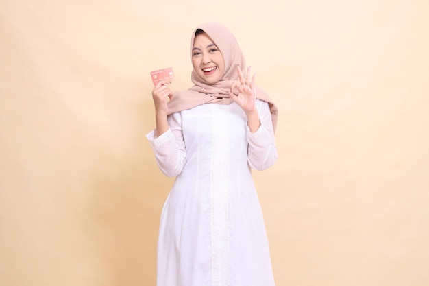 Индонезийская мусульманка в хиджабе счастливо улыбается, держа в руке дебетовую кредитную карту и жестикулируя окой.