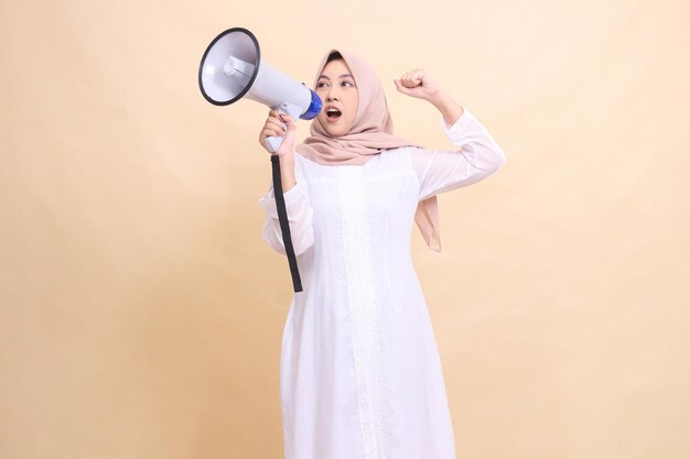 Индонезийская девушка в хиджабе, наклоненная направо, откровенно кричит с энтузиазмом, держа в руках громкоговоритель.