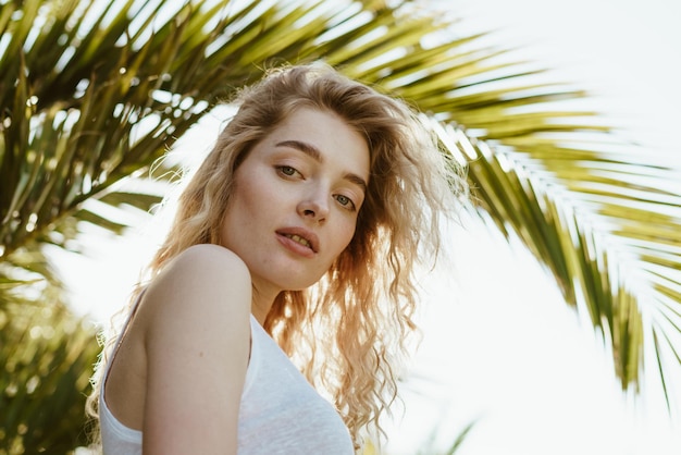 Girl inquiringly looks at the camera from top to bottom, standing under a palm branch