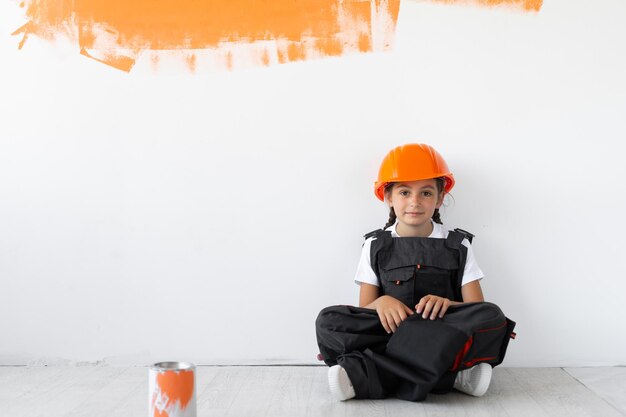 The girl is dressed in overalls and an orange protective helmet on her head. sitting on the floor looking in front.