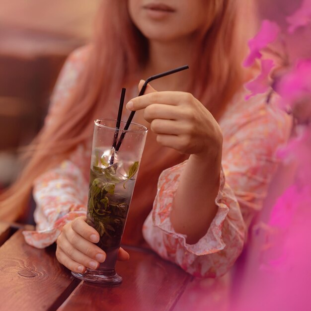 A girl is drinking a refreshing drink with mint through a straw from a glass A customer at a table in a summer cafe Woman's hands close up Pink colors