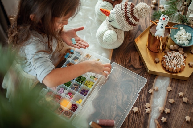 女の子はクリスマスの準備をしていて、ボタンで編まれた雪だるまを飾ります。新年の装飾のコンセプト。ジンジャーブレッドクッキーがテーブルの上にあります。ココアとクリスマスツリーの枝。