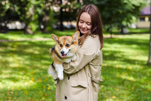 한 소녀가 그녀의 팔에 코기 개를 안고 있습니다. 녹지가 가득한 공원에서 여주인과 그녀의 애완 동물 산책