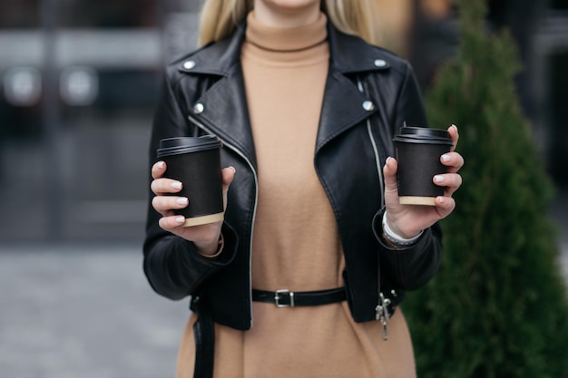 女の子は店の背景に女性の手で彼女の紙コップのコーヒーの隣に2つの紙コップを持っていますホットドリンク手はコーヒーの紙コップを持っていますコーヒー