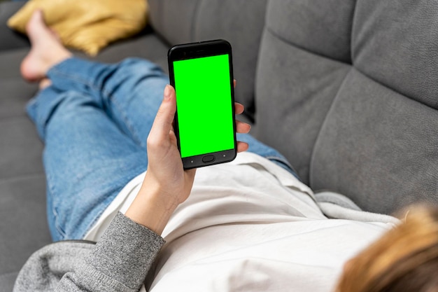 Girl is lying on the couch holding a smartphone with a green screen