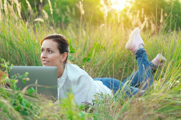 The girl is lying on the grass with a laptop in a remote corner of the city park. The concept of online learning and business