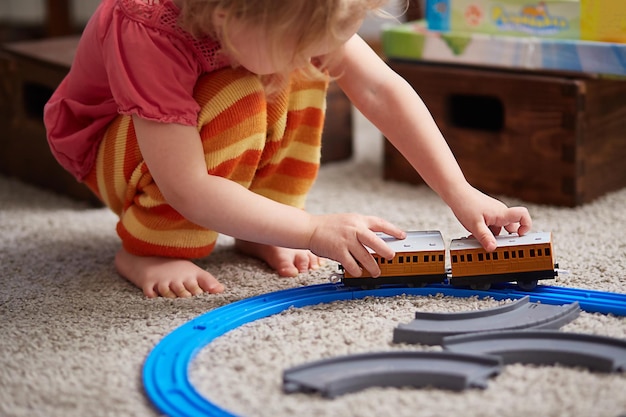 女の子は電車で遊んでいます。就学前および幼稚園の子供のための知育おもちゃ。女の子が家や幼稚園でおもちゃの鉄道を作っています。