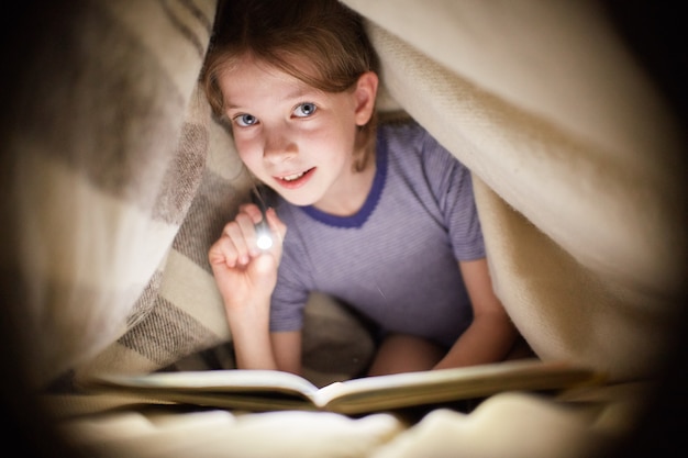 Girl is reading a book under a blanket with a flashlight in a dark room at night