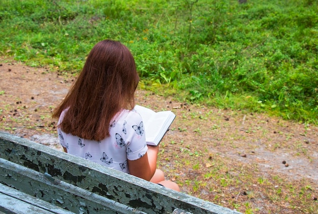 The girl is reading a book in the woods on the bench