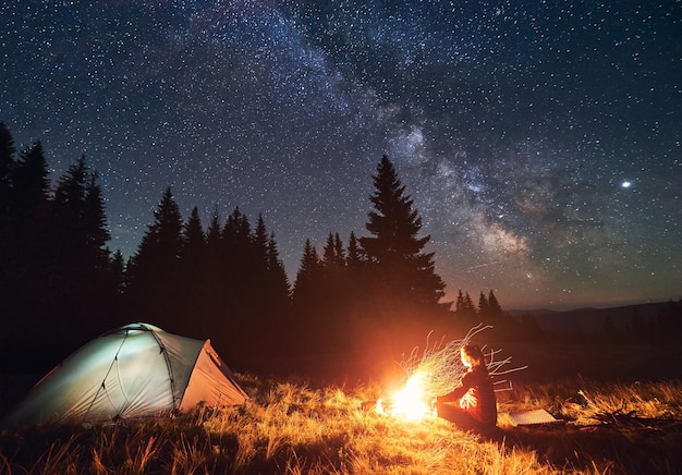 Girl is sitting by the fire on background of tent and spruce forest under starry sky on which milky way is visible