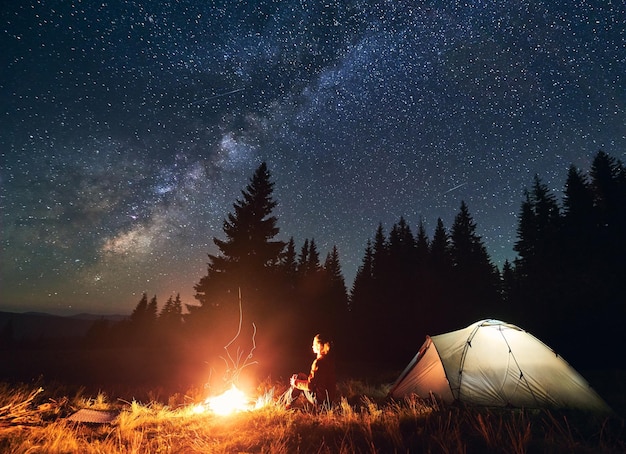 Girl is sitting by the fire on background of tent and spruce forest under starry sky on which milky way is visible