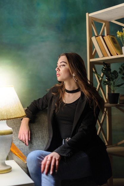 The girl is sitting in a chair A young girl near a floor lamp and a bookshelf