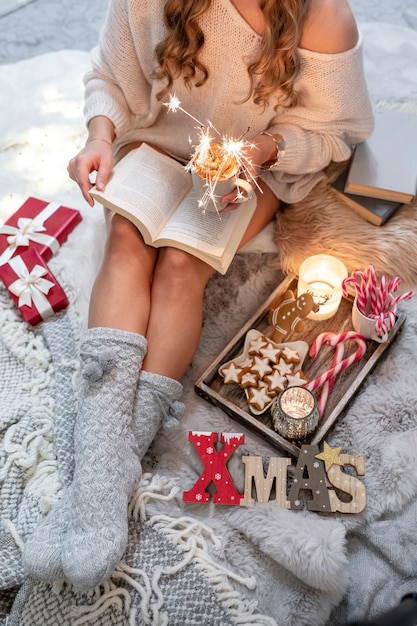 The girl is sitting in a christmas atmosphere, drinking a hot drink and reading a book.