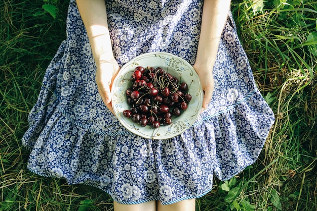 소녀는 파란색 빈티지 드레스를 입고 잔디에 앉아 있다. 여자는 체리를 들고 있다. 소박한 여름 과일이 평평하게 놓여 있습니다. 건강한 채식 생태 식품 라이프 스타일 개념입니다.