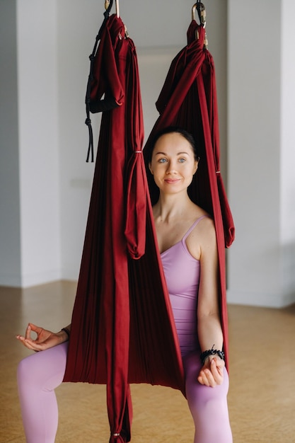 The girl is sitting in a hanging hammock and doing yoga in the gym