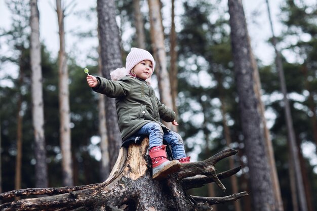 A girl is sitting on a stump in the woods with a Chupa Chups