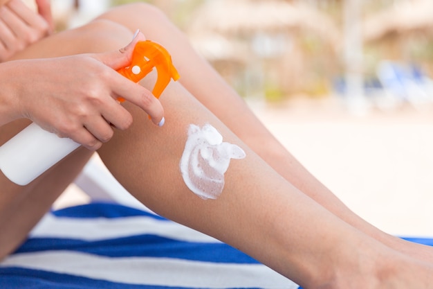 Girl is sitting on the sunbed and applying sun cream oh her leg at the beach.