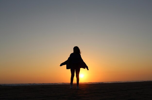 Foto una ragazza è in piedi su una spiaggia con il sole che tramonta dietro di lei