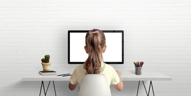 Girl is studying and working on the computer The concept of using modern technologies for children Work desk in the room with books and pens