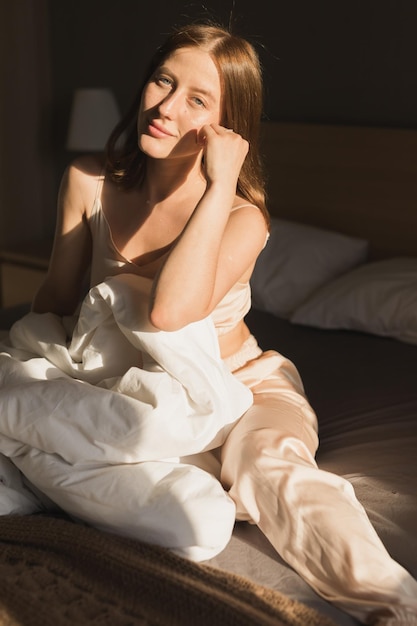 Girl is waking up in morning stretches in bed and sun shines from the window Happy young woman greets new sunny day