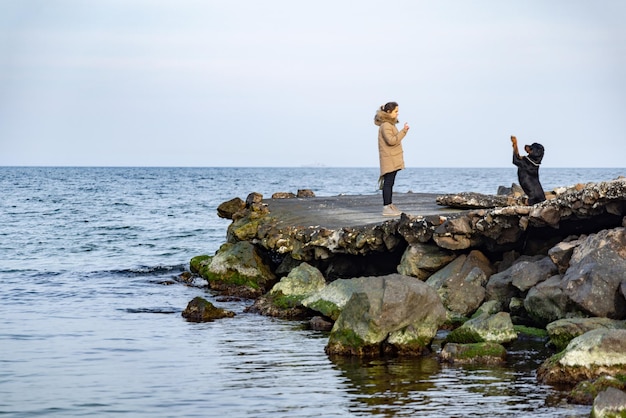 A girl in a jacket walks on the coast of the sea with a Rottweiler dog