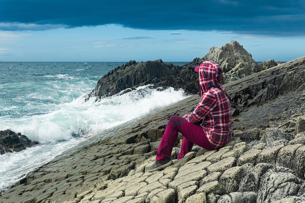 La ragazza con una giacca con cappuccio si siede sulla riva di un mare in tempesta