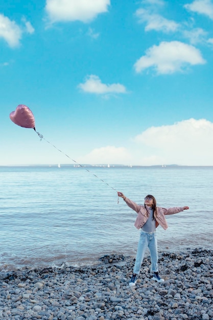 A girl in jeans and a pink jacket holding a balloon on the seashore at sunset