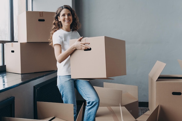 Girl in jeans and white tshirt is carrying cardboard box Happy attractive european woman packing things to move Packed boxes on the floor Relocation and moving to new apartment concept