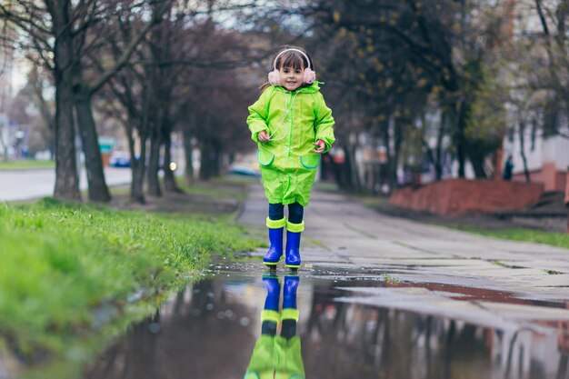 girl jumps on puddles in a green raincoat and blue boots