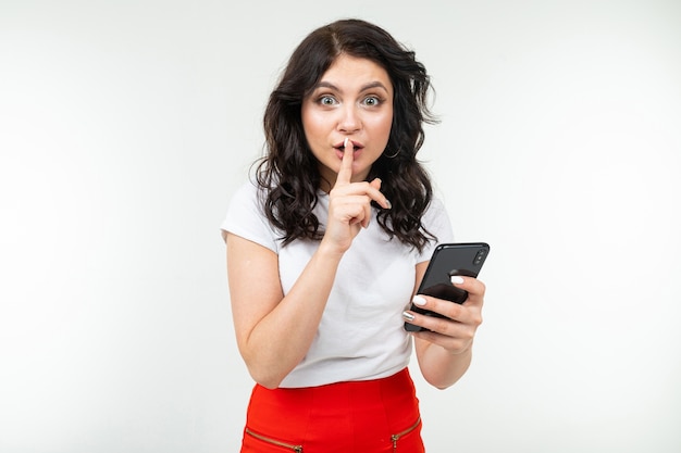 Girl keeps a secret while holding a finger at her mouth and a smartphone in her hand on a white background.