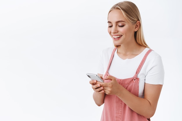 笑ってスマートフォンのディスプレイを見ている女の子は、面白いメッセージを受け取ります。オーバーオール、Tシャツ、ソーシャルメディアの閲覧、ビデオの視聴で携帯電話を使用する魅力的な金髪の現代白人女性