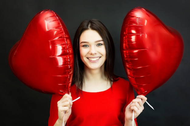 Girl laughs prettily holding balloons. A beautiful smiling brunette is holding two heart-shaped balloons near her face.