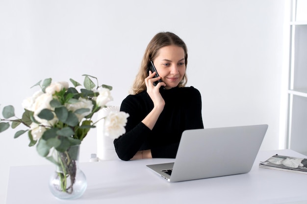 The girl laughs and talks on the phone in the office next to her laptop