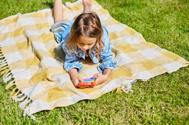 Girl lies on a blanket on grass outdoors and play pop it, kid hands playing with colorful pop It, fidget toy in the backyard of the house on a sunny summer day, summer time vacation.