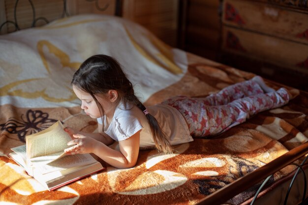 A girl lies on her stomach on a blanket on the bed and reads a book