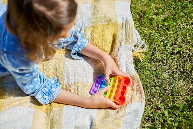 女の子は屋外の芝生の毛布の上に横たわって、それをポップで遊んだり、子供の手はカラフルなポップで遊んだり、晴れた夏の日、夏の休暇に家の裏庭でおもちゃをいじったりします。