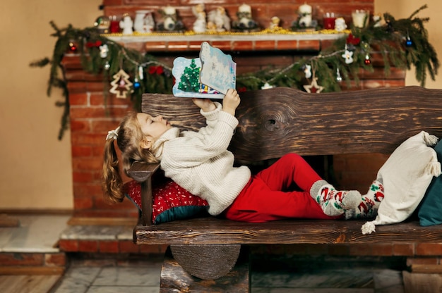 Girl lies with a book in her hands against the backdrop of a fireplace during the Christmas holidays