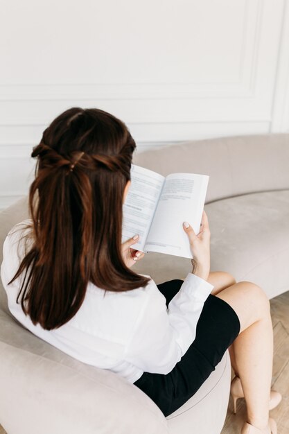 A girl in light clothes is sitting on the sofa and reading a book