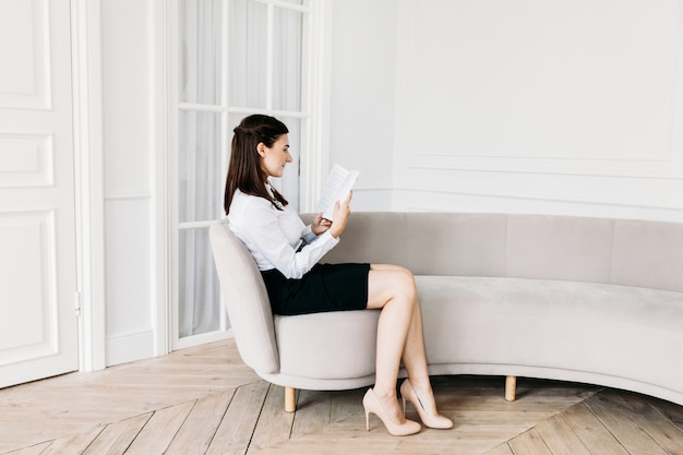 A girl in light clothes is sitting on the sofa and reading a book