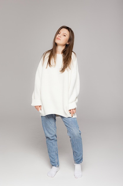 Девушка в светлых джинсах и белом свитере большого размера на сером фоне