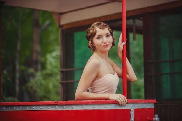 A girl in a light pink retro dress with a vintage hairstyle stands at the rail of the tram